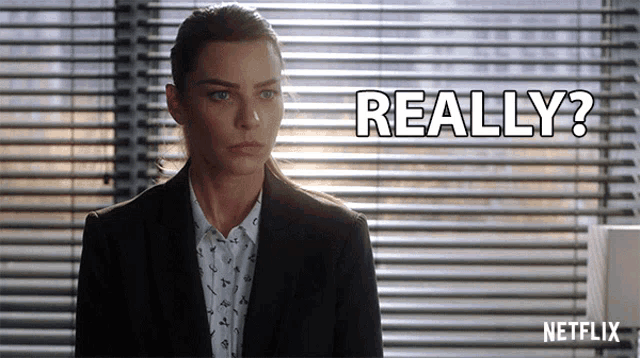 a woman in a suit is standing in front of a window with blinds and says " really "