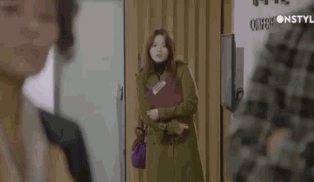 a woman in a trench coat is standing in a doorway holding a red folder .