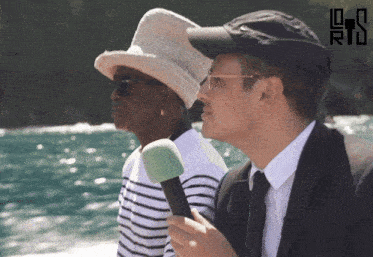 a man in a suit holds a microphone next to a woman wearing a hat