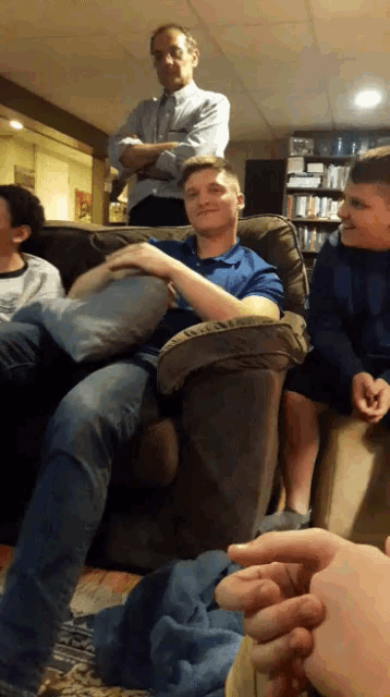 a man in a blue shirt sits on a couch with his arms crossed and a man in a suit stands behind him