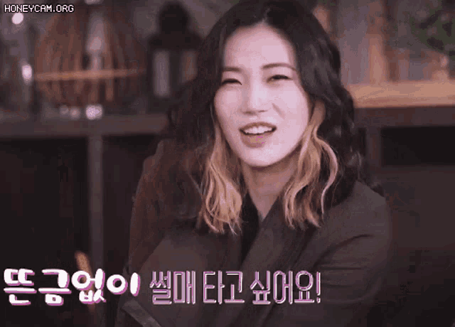 a woman is sitting on a couch and smiling with korean writing on the bottom of her face .