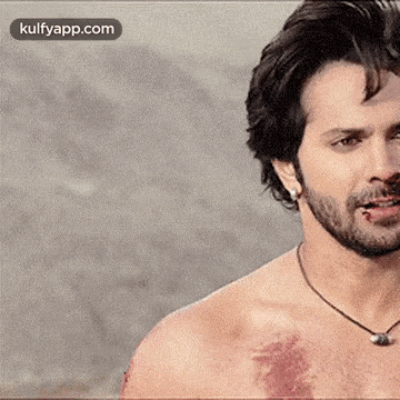 a shirtless man with a beard and a necklace is smoking a cigarette and looking at the camera .