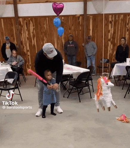 a man is holding a little girl while she plays with a bat and a piñata that looks like a unicorn