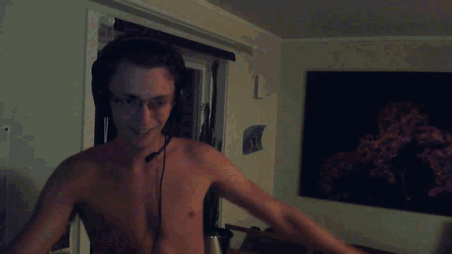 a shirtless man wearing headphones and glasses stands in front of a window