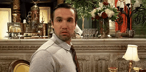 a man in a gray shirt and tie is sitting at a table in a restaurant with a vase of flowers in the background .