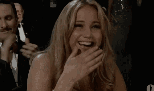 a woman is laughing while sitting in a dark room with a man in a tuxedo in the background .
