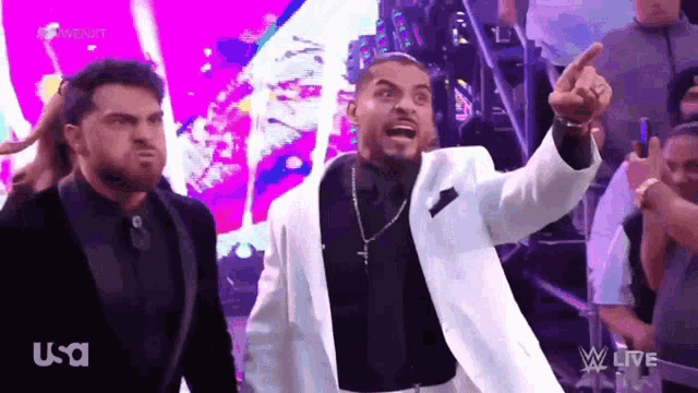 a man in a white suit and black shirt is pointing at something