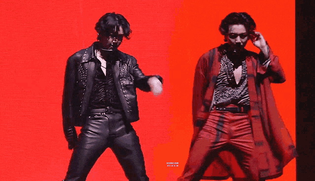 two men are dancing in front of a red background and one of them is wearing a jacket with studs