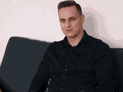a man in a black shirt is sitting on a couch and smiling