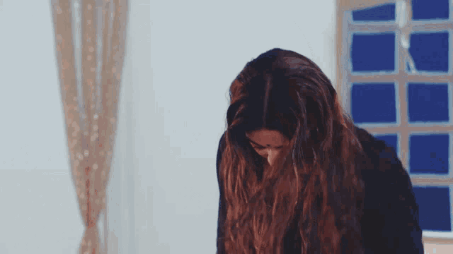 a woman with long hair looks down in front of a blue window