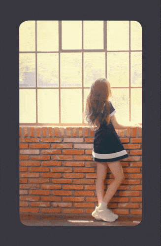 a little girl in a black and white dress leans against a brick wall looking out a window