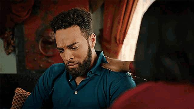a man with a beard is sitting on a couch while a woman holds his shoulders .