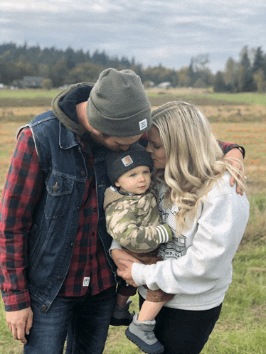 a man wearing a carhartt hat kisses a woman while holding a baby