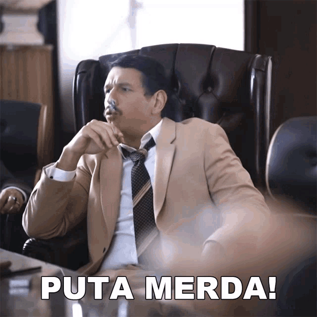 a man in a suit and tie is sitting in a chair with the words puta merda behind him