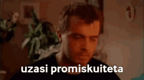 a man is sitting in a living room with a plant in the background and the words uzasi promiskuiteta written on his face .