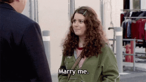 a woman in a green jacket is talking to a man in a suit and saying `` marry me '' .