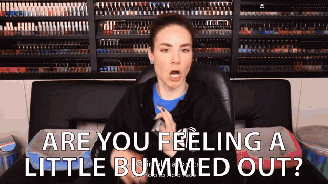 a woman is sitting in front of a wall of nail polish shelves and says " are you feeling a little bummed out "