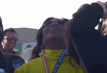 a woman wearing sunglasses and a lanyard that says new shepard is looking up at the sky .