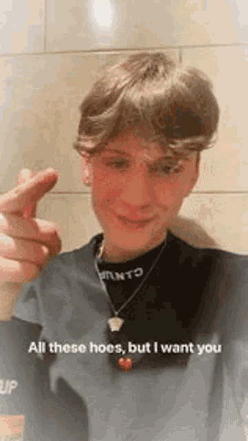 a young man wearing a black shirt and a necklace is making a heart with his finger .