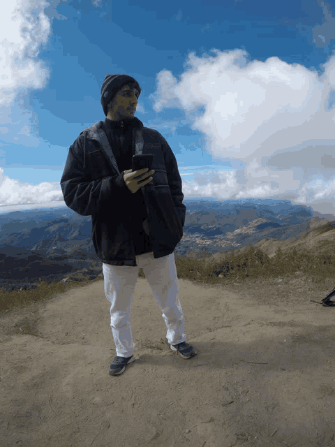 a man standing on a dirt road holding a cell phone with mountains in the background