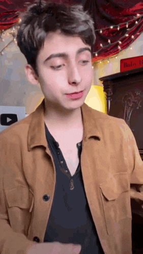 a young man wearing a tan jacket and a black shirt is standing in a room .