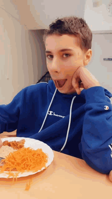 a young boy wearing a blue champion hoodie is eating a plate of food