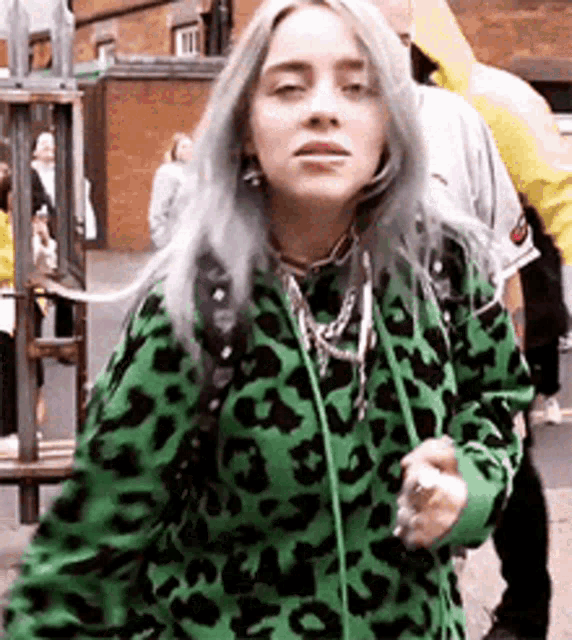 billie eilish is wearing a green and black leopard print hoodie while walking down the street .