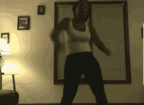 a woman in a white tank top and black pants is dancing in a dark room
