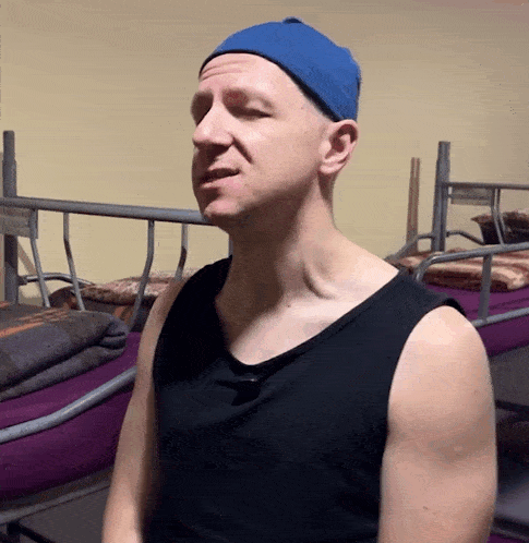 a man wearing a blue hat and a black tank top is standing in front of bunk beds