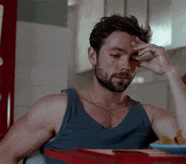 a man with a beard wearing a blue tank top is sitting at a table with his hand on his forehead