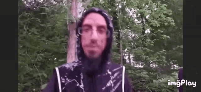 a man in a hooded jacket and glasses is standing in the woods .