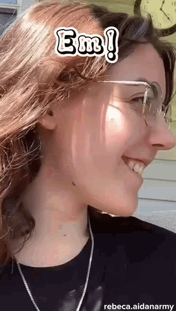 a woman wearing glasses and a necklace is smiling and says em