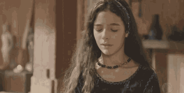 a young girl wearing a necklace and a headband is sitting in a room with her eyes closed .