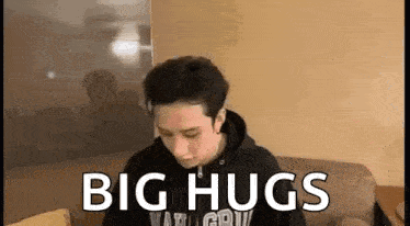 a man in a black hoodie is sitting on a couch with the words `` big hugs '' written on his face .