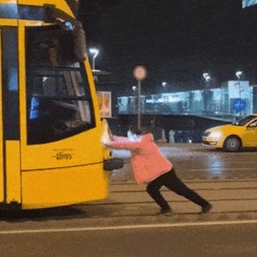 a woman in a pink jacket is pushing a yellow bus on a city street .