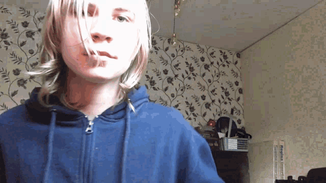 a young man wearing a blue hoodie stands in front of a wall with flowers on it