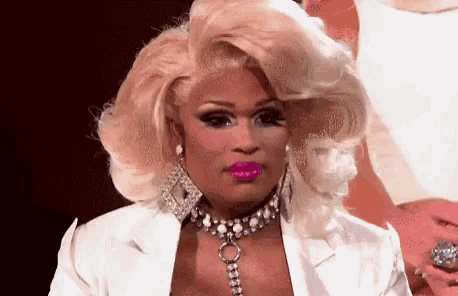 a drag queen with blonde hair and pink lipstick is wearing a white jacket and a choker necklace .