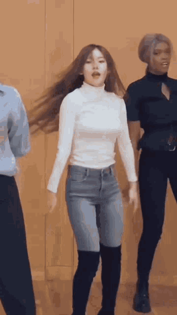 a woman in a white turtleneck and jeans is dancing in front of a wall .