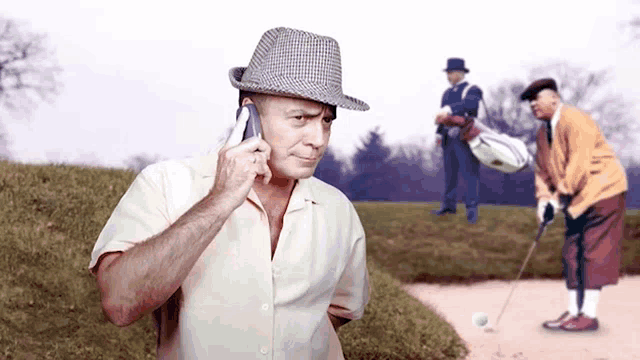 a man in a hat is talking on a cell phone while another man plays golf