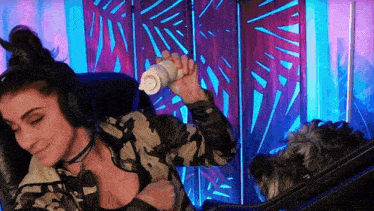 a woman in a camo jacket is holding a bottle in her hand while a dog looks on .