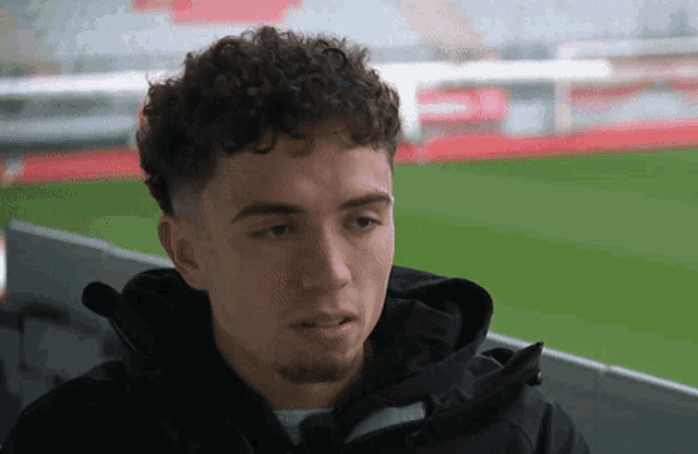 a young man with curly hair wearing a black jacket stands in front of a soccer field