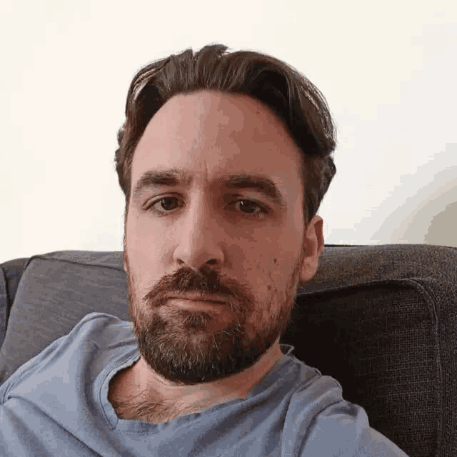 a man with a beard wearing a blue shirt is sitting on a couch