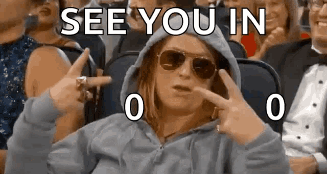 a woman wearing a hoodie and sunglasses is sitting in a crowd of people and making a peace sign .