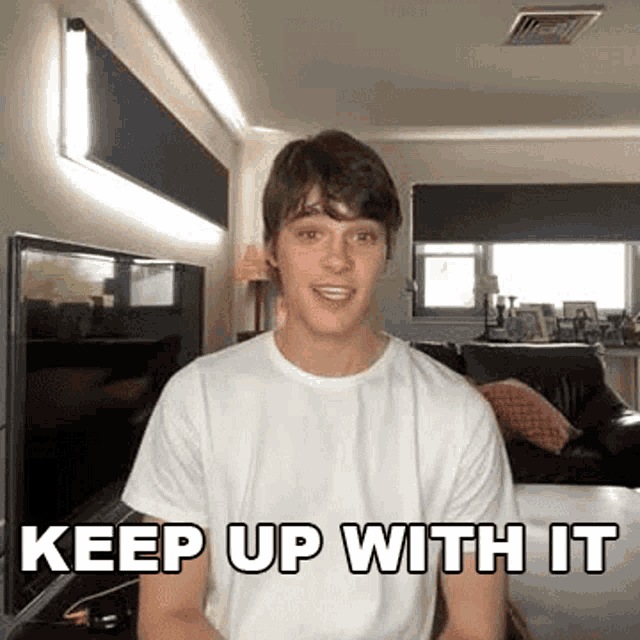 a young man in a white t-shirt is sitting in a living room with the words `` keep up with it '' .
