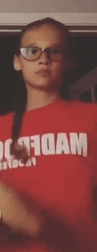 a young girl wearing glasses and a red shirt with the word foam on it is standing in front of a window .