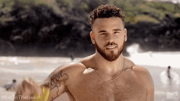 a shirtless man with a beard and tattoos on his arm is standing on a beach .