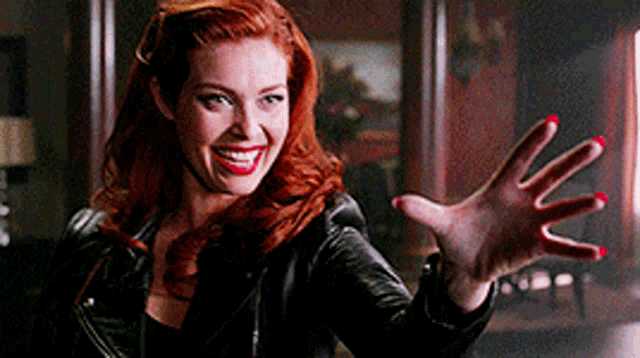 a woman with red hair and red nails is wearing a black leather jacket and waving her hand .
