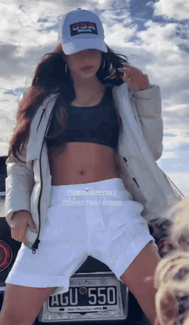 a woman in a crop top and white shorts is standing in front of a car with a license plate that says agu 550