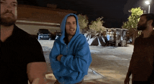 a man in a blue hoodie is standing with his arms crossed in a parking lot