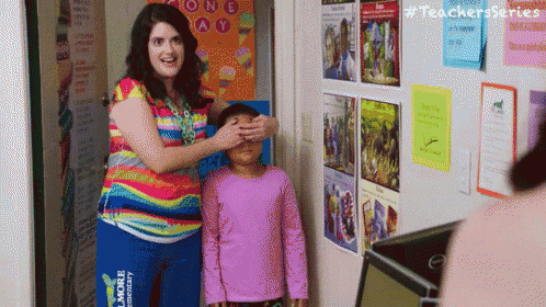 a woman is covering a child 's eyes in a classroom with posters on the wall .